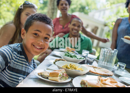 Una riunione di famiglia, uomini, donne e bambini intorno a un tavolo in un giardino in estate. Un ragazzo sorridente in primo piano. Foto Stock