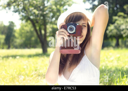 Giovane donna graziosa in posa con la vecchia telecamera cinematografica nel parco d'estate. Ragazza nel cappello bianco fotografia con fotocamera manuale Foto Stock