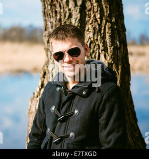 Giovane uomo bello appoggiato contro un albero dal fiume In autunno il giorno, sorridente nella fotocamera. Uno stile informale - Giacca, occhiali da sole Foto Stock
