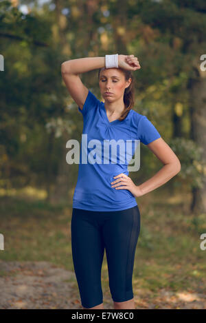 Donna atleta tergi sudore dalla sua fronte sul suo cinturino da polso come si ferma durante la sua formazione esercizi su una pista forestale Foto Stock