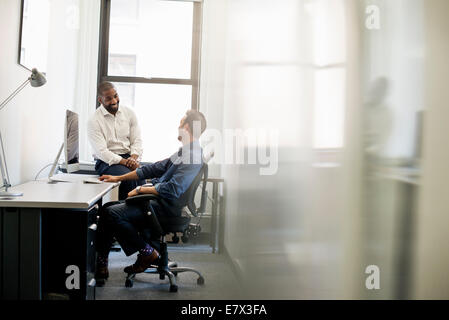 Vita in ufficio. Un uomo Pendente ritornare in una sedia da ufficio a parlare con un collega seduto sul bordo della scrivania. Foto Stock
