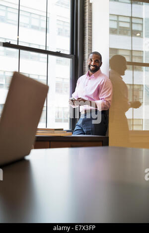 Vita in ufficio. Un uomo in piedi da una finestra in un ufficio controllo il suo smart phone. Foto Stock