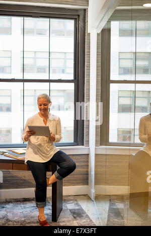 Vita in Ufficio.Una donna seduta sul bordo della sua scrivania utilizzando una tavoletta digitale. Foto Stock