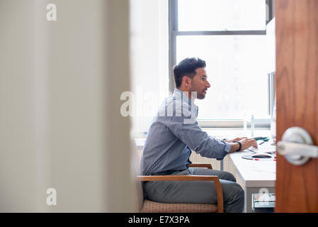 Vita in ufficio. Un uomo seduto a una scrivania utilizzando un computer, guardando attentamente lo schermo. Foto Stock