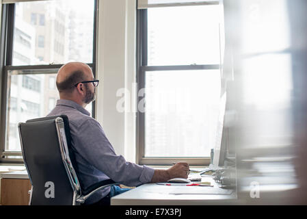 Vita in ufficio. Un uomo seduto a una scrivania utilizzando un computer, guardando attentamente lo schermo. Foto Stock