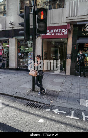 Una giovane ragazza si appoggia contro un semaforo con il suo telefono cellulare in possesso di una borsa a mano mentre in attesa di attraversare la strada a Londra Foto Stock