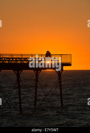 Due persone stand su Aberystwyth pier stagliano da il sole tramontare all'orizzonte. Foto Stock