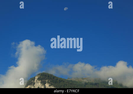 Una fotografia della luna in una mattina blu cielo sopra una montagna con una copertura di nuvole. Foto Stock