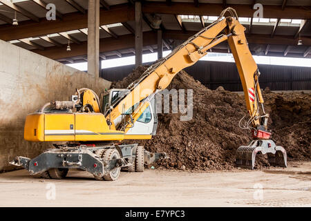 Per impieghi pesanti escavatore facendo movimento terra undercover parcheggiata in un magazzino aperto davanti a un enorme mucchio di terra bruna per constr Foto Stock