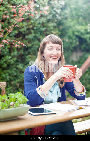 Donna seduta all aperto con tablet pc, tenendo la tazza e sorridente Foto Stock