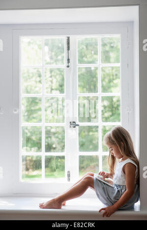 Una giovane ragazza seduta in corrispondenza di una finestra sede utilizzando una tavoletta digitale. Foto Stock