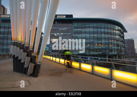 La sede principale della BBC per la trasmissione televisiva a nord a MediaCity, Media City, Salford Quays con uno jogger che attraversa il ponte pedonale MediaCityUK link illuminato all'alba. Foto Stock