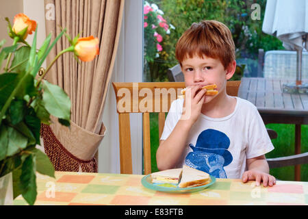 Un giovane ragazzo seduto a tavola a mangiare un panino Foto Stock