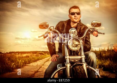 Biker uomo che indossa una giacca di pelle e occhiali da sole seduto sulla sua moto guardando il tramonto. Foto Stock