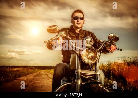 Biker uomo che indossa una giacca di pelle e occhiali da sole seduto sulla sua moto guardando il tramonto. Foto Stock