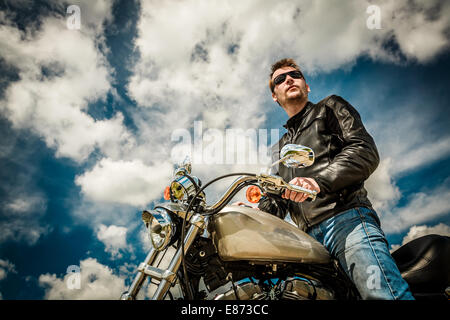 Biker uomo che indossa una giacca di pelle e occhiali da sole seduto sulla sua moto guardando il tramonto. Foto Stock