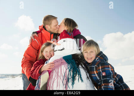 Ragazzo e una ragazza in piedi con pupazzo di neve, genitori baciare in background, Baviera, Germania Foto Stock