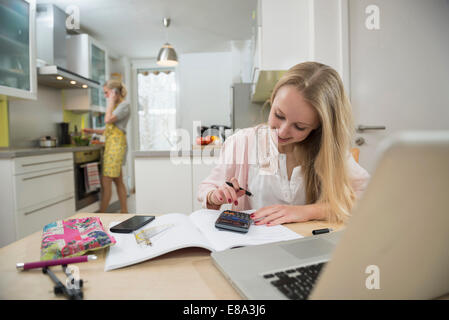 Ragazza adolescente facendo compiti a casa mentre sua madre a preparare il pranzo, sorridente Foto Stock