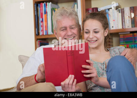 Nonno e nipote leggendo un libro sul divano nel soggiorno, sorridente Foto Stock