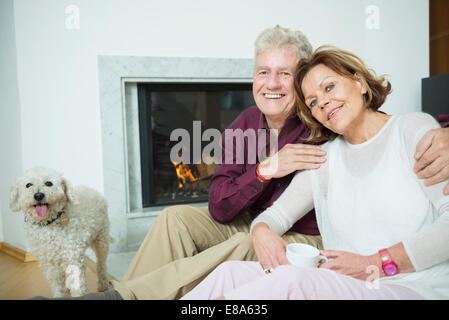 Ritratto di coppia senior con il cane seduto di fronte al caminetto, sorridente Foto Stock
