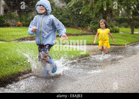 Ragazzo e sorella di indossare stivali di gomma in esecuzione e sguazzare nella pozza di pioggia Foto Stock