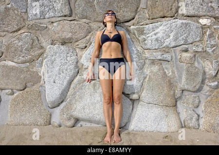 Una donna nella sua metà degli anni quaranta sunbathes mentre giace contro una parete in Cornwall, Regno Unito Foto Stock