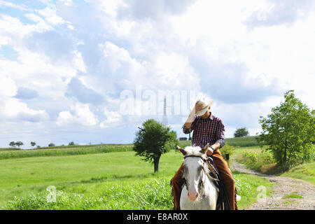 Giovane uomo regolando il cappello da cowboy a cavallo sulla strada rurale Foto Stock