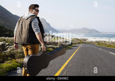 Vista posteriore del giovane uomo a camminare sulla strada costiera che porta la chitarra caso, Cape Town, Western Cape, Sud Africa Foto Stock