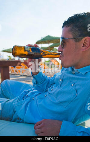 Ritratto verticale di un giovane attraente uomo marocchino di bere una birra su di una spiaggia. Foto Stock