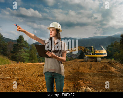 Ritratto di giovane donna caucasica architetto in piedi sul sito in costruzione, tenendo appunti, puntando a qualcosa di Foto Stock