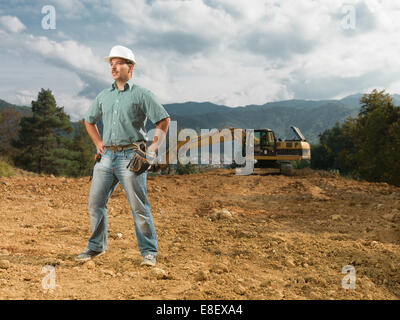Giovani caucasici ingegnere maschio in piedi sul sito in costruzione, con escavatore in background Foto Stock