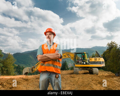 Vista anteriore dell'ingegnere in piedi sul cantiere aperto con escavatore in background Foto Stock