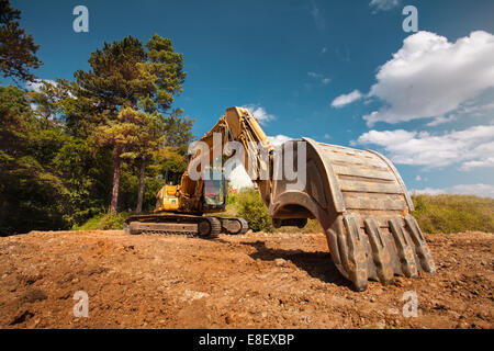 Vista prospettica di escavatore sul nuovo sito in costruzione con alberi e cielo blu in background Foto Stock