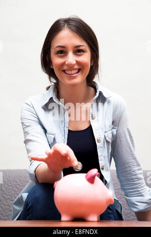 Ritratto di una giovane donna sorridente che inserisce una moneta in una banca piggy Foto Stock