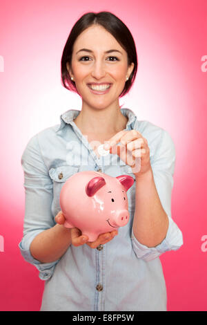 Ritratto di una giovane donna sorridente che inserisce una moneta in una banca piggy Foto Stock