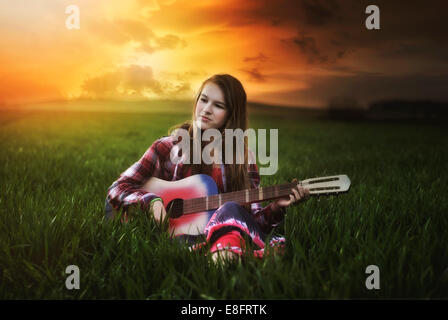 Sorridente ragazza seduta in un prato al tramonto suonando la chitarra, Polonia Foto Stock