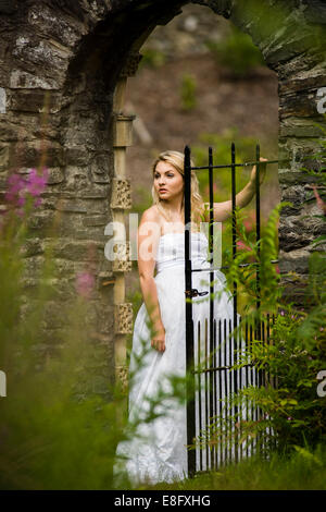Una giovane donna bionda ragazza modello 'sposa indossa un bianco Palla abito abiti da sposa in piedi in un gateway in un muro di pietra nel giardino Foto Stock