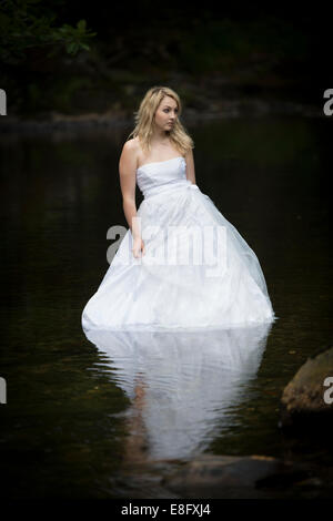 "Cestino il vestito": una giovane donna bionda ragazza modello sposa che indossa la sfera bianca abito abiti da sposa in piedi in lento fiume che scorre Foto Stock