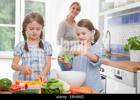 Germania - Monaco, ragazze (4-7) tritare verdure in cucina, madre in piedi in background Foto Stock