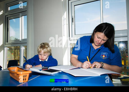 Bambino in pre-scuola di classe con Learning Support Assistant (LSA), Oxfordshire, Regno Unito iscritto in un libro di scuola Foto Stock