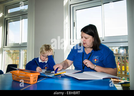 Bambino in pre-scuola di classe con Learning Support Assistant (LSA), Oxfordshire, Regno Unito iscritto in un libro di scuola Foto Stock