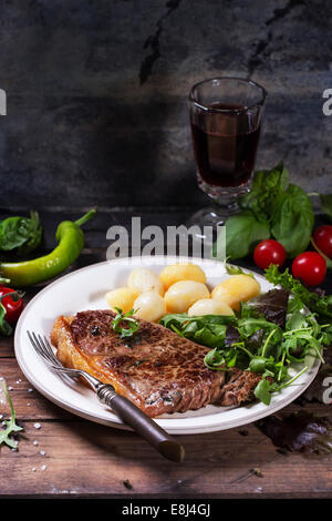 Bistecca alla griglia con patate e insalata verde su bianco piastra ceramica e vetro vintage vino rosso con sopra il vecchio tavolo in legno.