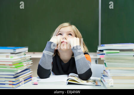 La scolaretta, 9 anni, guardando annoiato, seduto tra due pile di libri di fronte a una lavagna Foto Stock