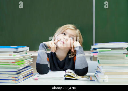 La scolaretta, 9 anni, guardando pensieroso, seduto tra due pile di libri di fronte a una lavagna Foto Stock