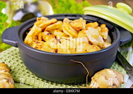 Topinambur arrostito in una teglia con un coperchio, tuberi freschi su un tovagliolo, prezzemolo, olio vegetale su tavole di legno b Foto Stock