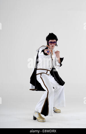 Uomo vestito in abiti fantasiosi, costume da commedia come Elvis, in pieno su degli anni settanta jump suit, con razzi, parrucca e occhiali da sole e stivali di piattaforma Foto Stock