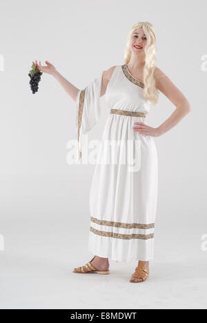 Ragazza vestita in costume costume da commedia come un romano / edyptian lady in un mantello bianco, parrucca bionda, sandali e uva Foto Stock