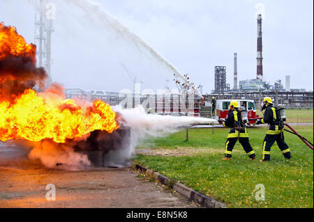 Due vigili del fuoco in uniforme di protezione e casco di estinguere un incendio con schiuma contro lo sfondo della raffineria di petrolio. Foto Stock