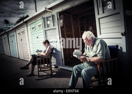 L uomo e la donna seduta su sedili di legno la lettura al di fuori di una fila di cabine sulla spiaggia, sul lungomare a Lyme Regis. Foto Stock