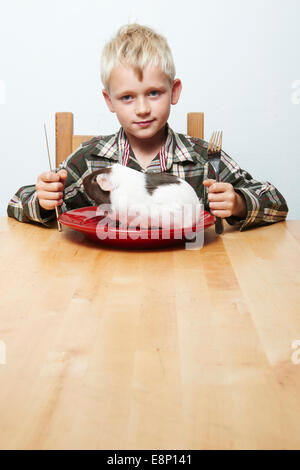 Bambino biondo ragazzo seduto al tavolo sempre pronto a mangiare con posate cavia (carne cruda) seduto su una targhetta rossa Foto Stock